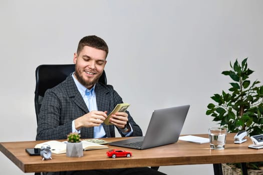 young bearded businessman working on laptop and holding money while sitting on chair at desk.