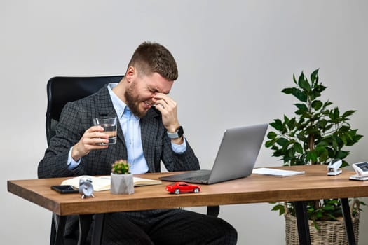 bearded man sitting on chair at desk and suffering from headache at workplace, using laptop