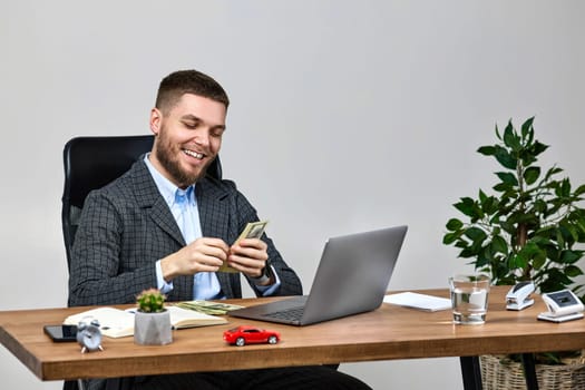 young bearded businessman working on laptop and holding money while sitting on chair at desk.