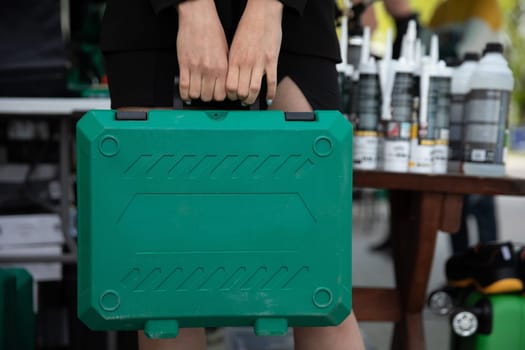 A woman's hands hold a green suitcase with power tools by the handle. Scene taken on a sunny day outside.