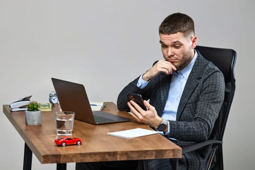 young bearded businessman sitting on chair at desk and holding phone, using laptop