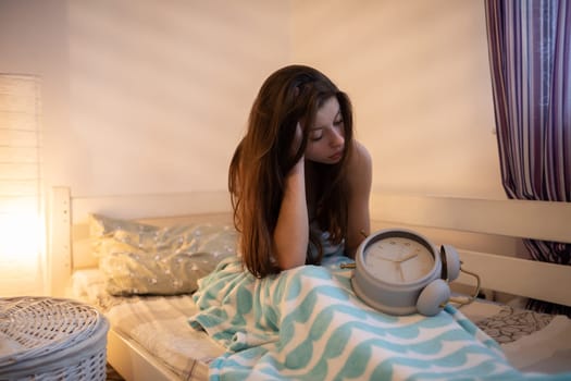 The girl sits offended with the alarm clock in her lap. The light of a lamp on the wall.