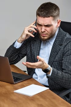 Upset angry bearded businessman sitting on chair at desk and talking on the phone, using laptop