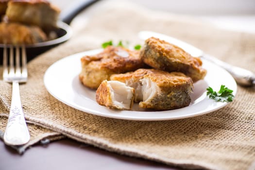 A piece of hake fish fried in a crispy crust, in a plate on a wooden table.