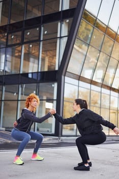 Does squats. Two female friends with sportive bodies have fitness day outdoors.