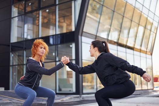 Does squats. Two female friends with sportive bodies have fitness day outdoors.