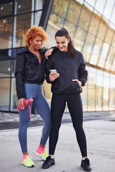 Holds phone in hands. Two female friends with sportive bodies have fitness day outdoors.