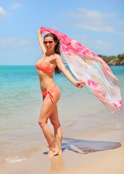 Young athletic slim woman in orange bikini and sunglasses stands on the beach, holding scarf behind her that is waving in the wind, azure sea background.