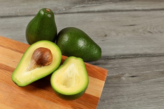 Avocado cut in half brown seed visible, wooden chopping board under, two whole green pears next to it.