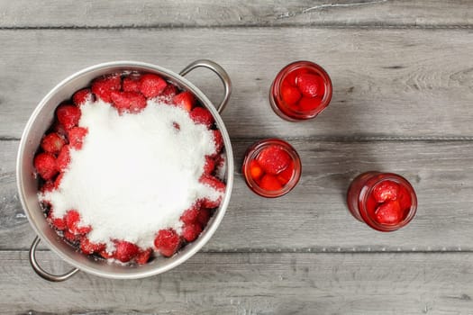 Tabletop view, large steel pot full of strawberries covered with crystal sugar, three bottles of strawberry in syrup next to it on gray wood desk. Homemade compote preparation.