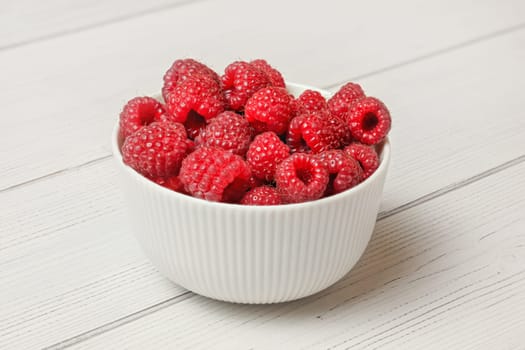 Small white ceramic bowl full of freshly picked raspberries, light boards desk under.