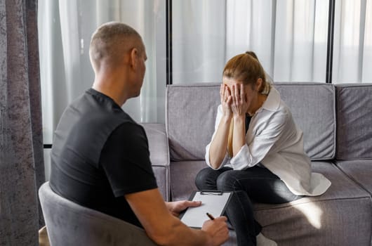 A male psychologist consults a female patient, makes notes. A woman emotionally talks about herself, covering her face with her hands.