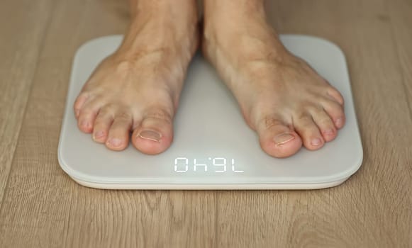 Man weighing himself - male bare feet stepping on white digital floor scales at home: close up view. Measuring weight, control, wellness and diet concept.
