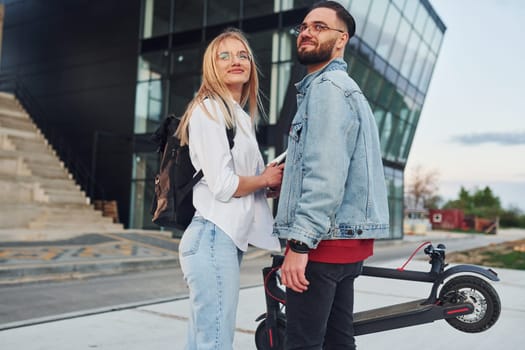 With electric schooter. Young stylish man with woman in casual clothes outdoors near business building together. Conception of friendship or relationships.