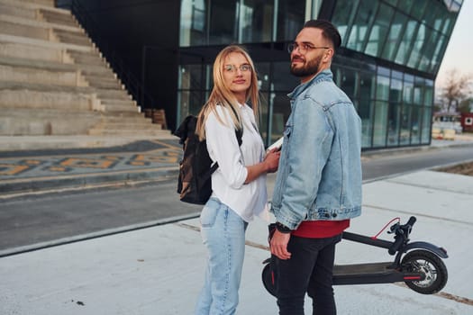 With electric schooter. Young stylish man with woman in casual clothes outdoors near business building together. Conception of friendship or relationships.