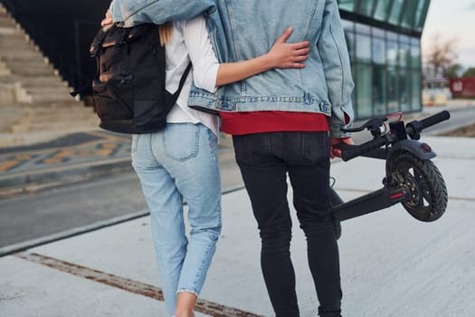 With electric schooter. Young stylish man with woman in casual clothes outdoors near business building together. Conception of friendship or relationships.