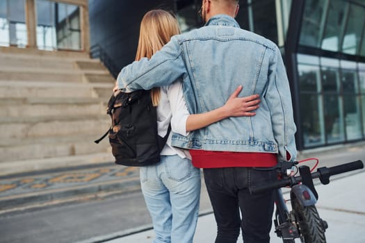 With electric schooter. Young stylish man with woman in casual clothes outdoors near business building together. Conception of friendship or relationships.