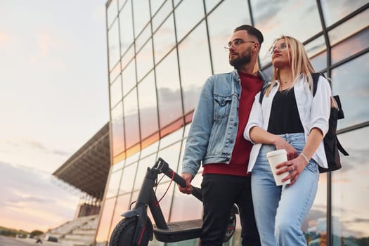 With electric schooter. Young stylish man with woman in casual clothes outdoors near business building together. Conception of friendship or relationships.