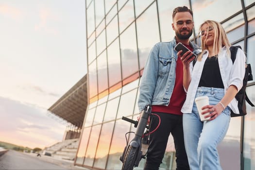 With electric schooter. Young stylish man with woman in casual clothes outdoors near business building together. Conception of friendship or relationships.