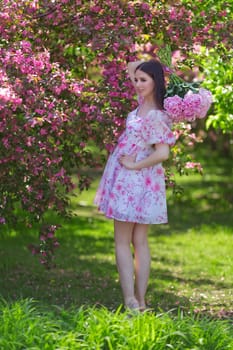 A beautiful happy girl with long hair, a brunette, in a light pink dress, holding a bouquet of large pink peonies behind her back, standing near pink flowering apple trees, in the garden on a sunny day in spring. Vertical