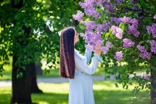 girl with long hair , in light blue dress stands nearby lilac flowers, in the garden, in sunny day. Close up. copy space