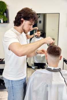 Professional barber does haircut for caucasian bearded man using comb and scissors at barber shop.