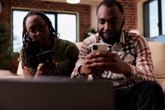 African american couple living together having issues not talking to each other spending time on smartphones sitting on couch. Boyfriend ignoring upset girlfriend after arguement in home living room.