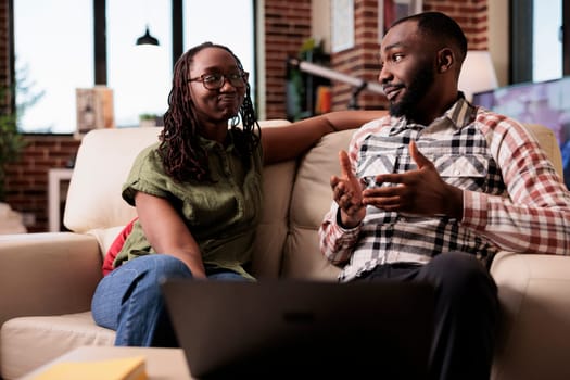 African american couple talking with each other while sitting on couch in home living room. Girlfriend looking discontented at boyfriend explaining and hand gesturing in front of table with laptop.