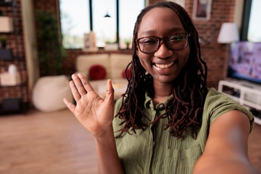 Happy african american woman waving hello in video call conference with friends in home living room. Casual smiling student doing greeting hand gesture at camera while talking with colleagues.