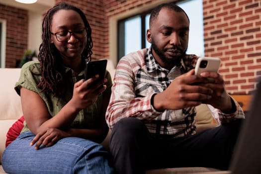 African american couple spending time looking at social media content on smartphones and ignoring each other on couch. Man and woman living together upset after arguement using mobile phones.