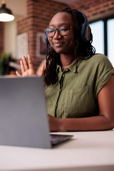 Portrait of african american student with wireless headphones doing hello hand gesture in video conference in living room. Smiling freelancer waving at laptop screen greeting client in internet call.