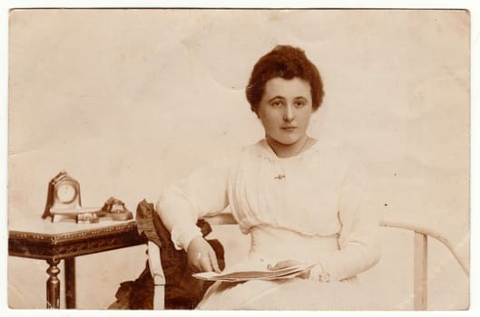 POZNAÅ  (POSEN), POLAND - CIRCA 1920s: Vintage photo shows a young woman. She wears a white dress. Historical table and an old world mantel clock are on the background. Retro black and white studio photography. Circa 1920s.