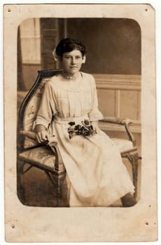 GERMANY - CIRCA 1920s: Vintage photo shows a young woman. She wears a white dress. She sits on the historical armchair and holds roses. Retro black and white studio photography.