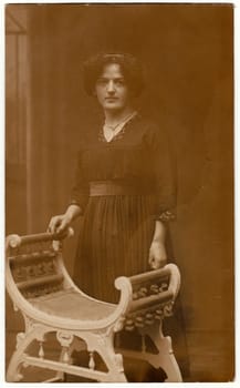 HAMBURG, GERMANY - APRIL, 1916: Vintage photo shows a young woman. She wears a black dress. She stands at a historical chair. Retro black and white studio photography. Circa 1920s.