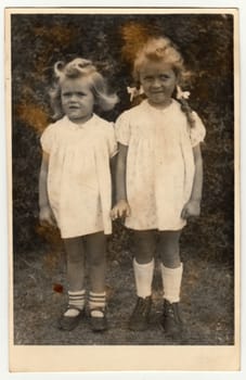 GERMANY - DECEMBER 23, 1946: Vintage photo shows two small girls siblings. They wear a white dresses. Retro black and white photography. Circa 1940s.