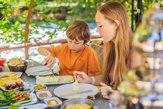 Mom and son eat Turkish breakfast. Turkish breakfast table. Pastries. Vegetables. Olives. Cheeses, fried eggs. Jams, tea in copper pot and tulip glasses. Wide composition.