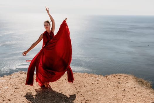 Side view a Young beautiful sensual woman in a red long dress posing on a rock high above the sea during sunrise. Girl on the nature on blue sky background. Fashion photo.