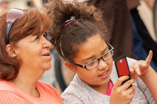 close-up of a teenager with a cell phone showing her mom and the mom looking elsewhere. High quality photo they both wear glasses and are on the street