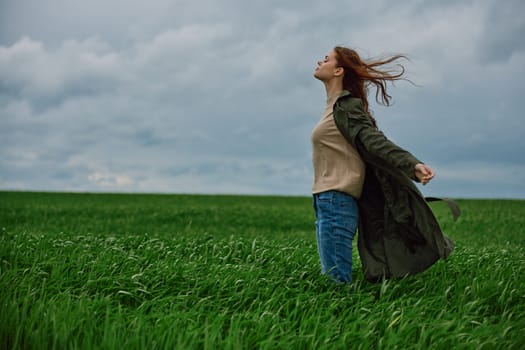 a red-haired woman in a long coat stands in a green field and the wind blows her hair. High quality photo