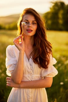 close portrait of a beautiful, laughing woman in a light dress holding a chamomile near her face during sunset, lit from the back. High quality photo