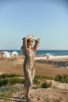 a slender, sophisticated woman in a beige dress stands on the sand in windy weather with her hands raised to her head. High quality photo