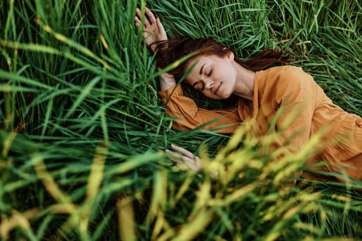 a close horizontal photo of a pleasant woman in a long orange dress resting lying in the tall grass with her eyes closed in sunny weather at sunset with her arms outstretched. Street photography, the theme of privacy with nature. High quality photo