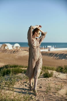 a slender, sophisticated woman in a beige dress stands on the sand in windy weather with her hands raised to her head. High quality photo