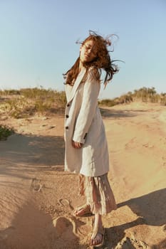 a woman poses standing on the sand in light summer clothes against the blue sky and the wind blows her hair on her face. High quality photo