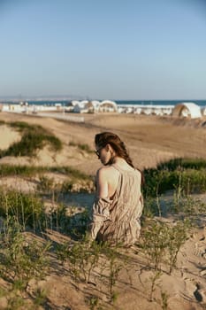red-haired woman posing sitting with her back to the camera in a beige dress on the sea coast during sunset. High quality photo