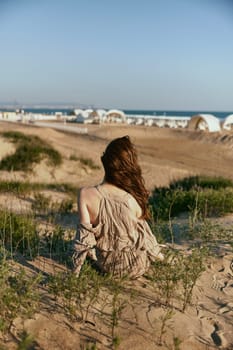 red-haired woman posing sitting with her back to the camera in a beige dress on the sea coast during sunset. High quality photo