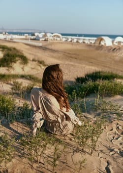 red-haired woman posing sitting with her back to the camera in a beige dress on the sea coast during sunset. High quality photo