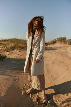 a woman poses standing on the sand in light summer clothes against the blue sky and the wind blows her hair on her face. High quality photo