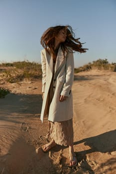 a woman poses standing on the sand in light summer clothes against the blue sky and the wind blows her hair on her face. High quality photo