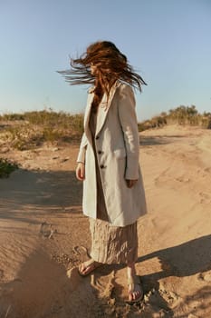a woman poses standing on the sand in light summer clothes against the blue sky and the wind blows her hair on her face. High quality photo